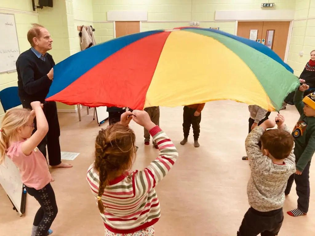 Children play with a colorful parachute while an adult leads the activity in a community room