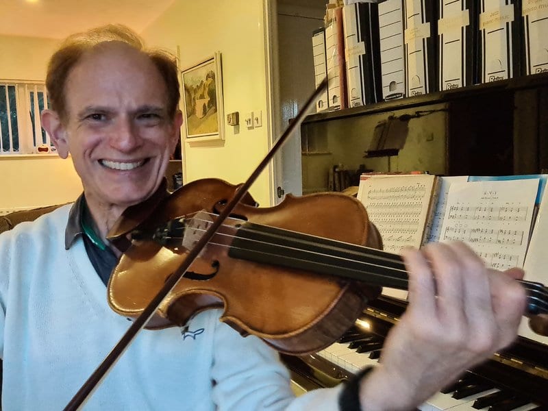 Smiling man playing a violin in front of a piano, surrounded by sheet music and files